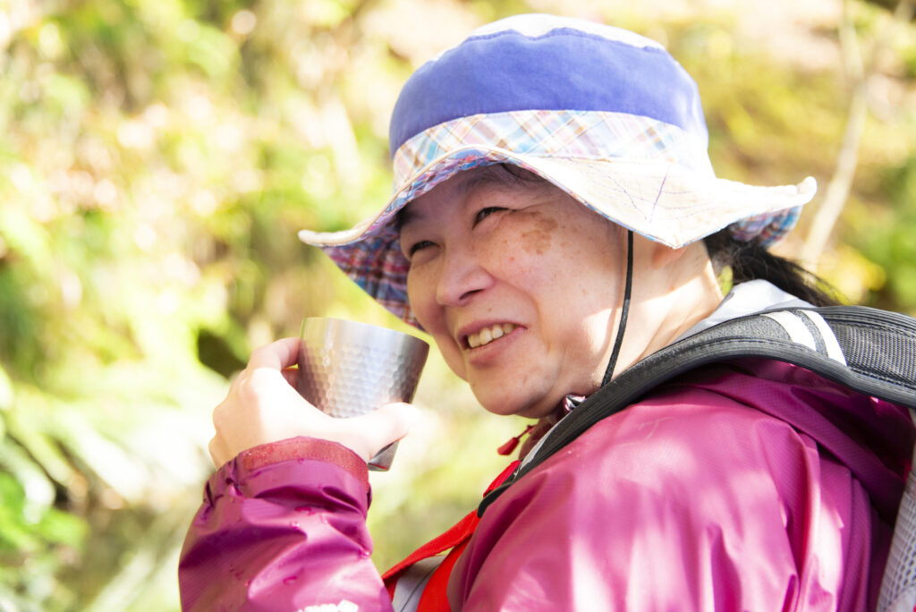 紅葉を背に、金属カップの温かい飲み物を笑顔で味わうさっちゃん