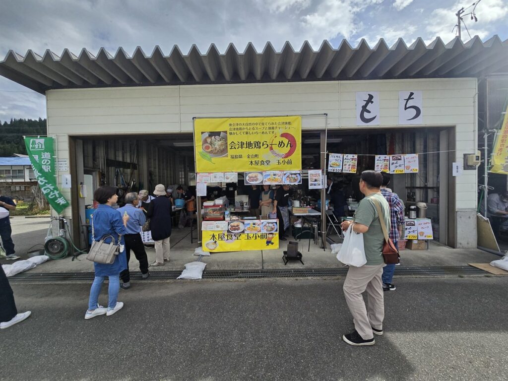 奥会津ごっつおまつりの出店。会津地鶏らーめんやお餅ののぼりが立ち、来場者が買い物をしている